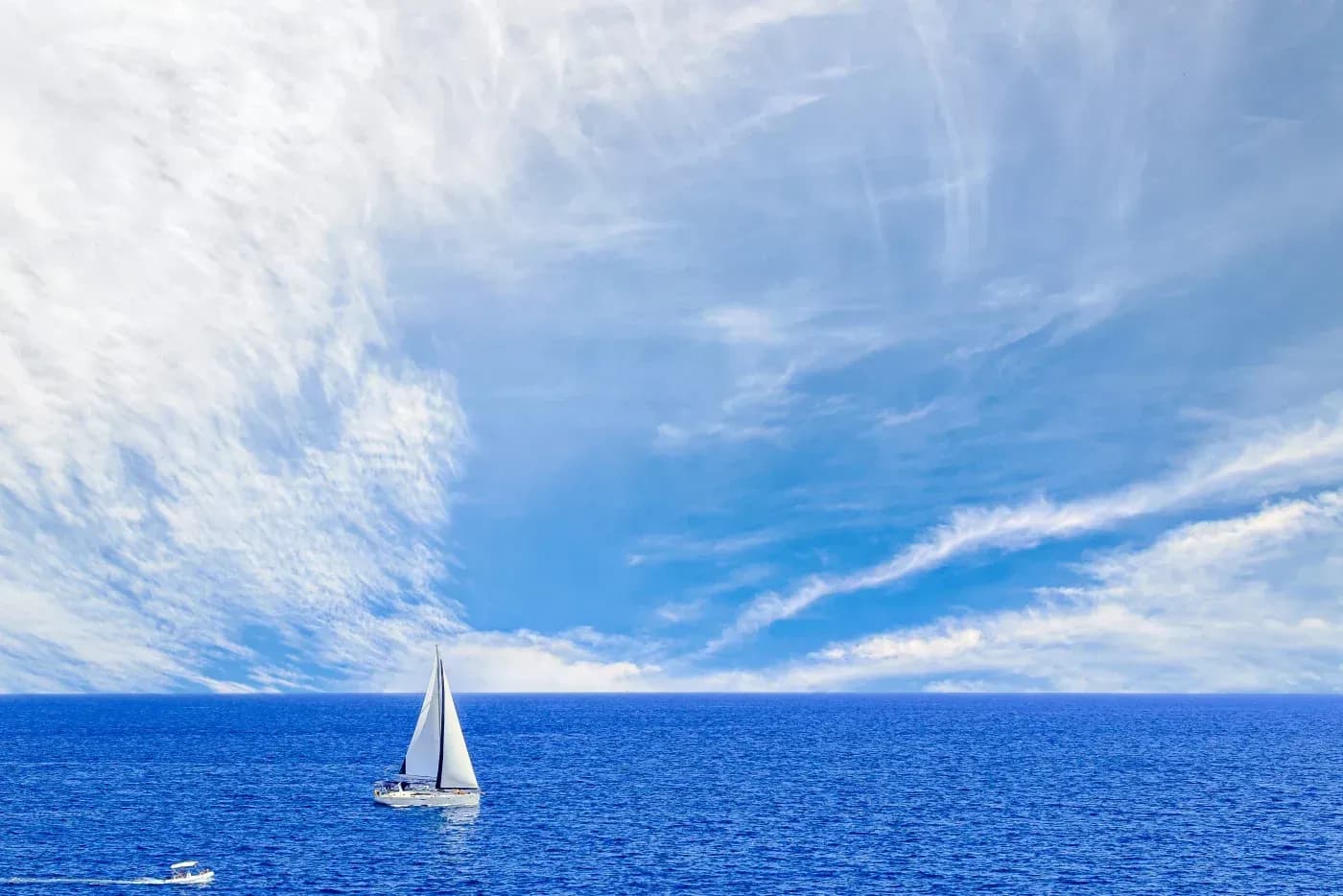 White sailboat cruising across a deep blue ocean beneath a bright sky filled with sweeping white clouds.