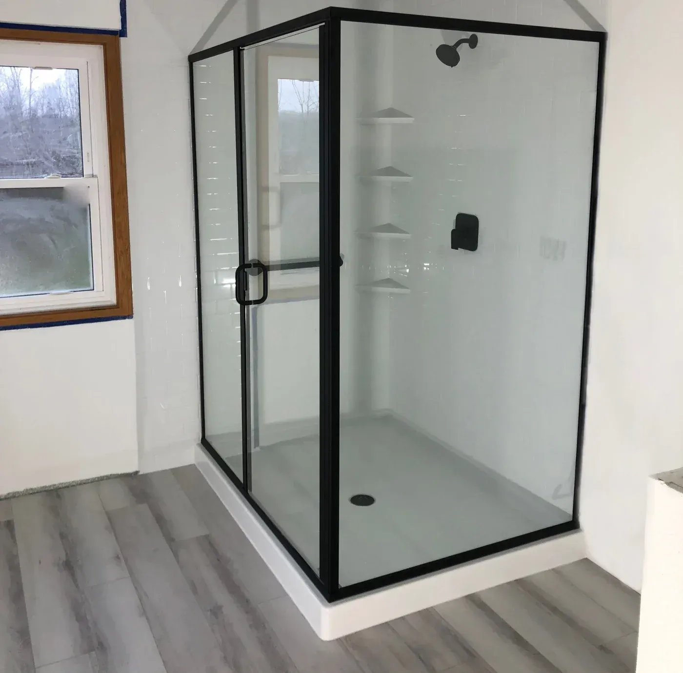 Modern black-framed corner shower enclosure with glass panels, overhead rainfall showerhead, and built-in shelving in a bathroom with wood-look flooring