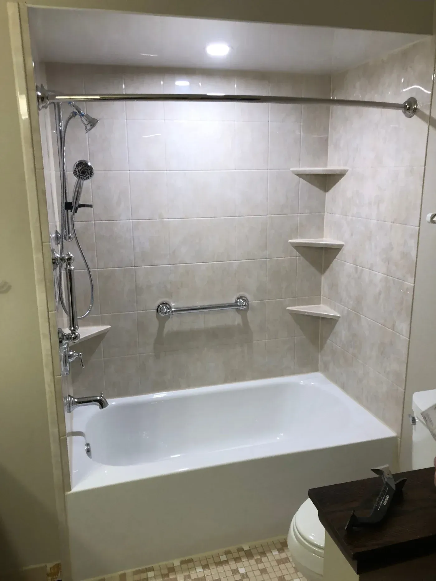 Bathroom shower enclosure with white tile surround, built-in corner shelves, handheld showerhead, grab bar, and white bathtub base