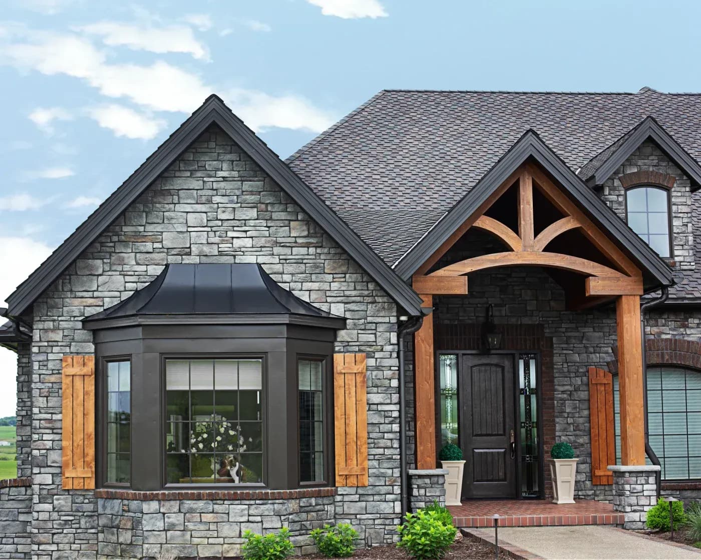 Luxury residential home with gray stone exterior, metal roof, black-framed bay window with wooden shutters, and timber-frame entry portico with landscaping