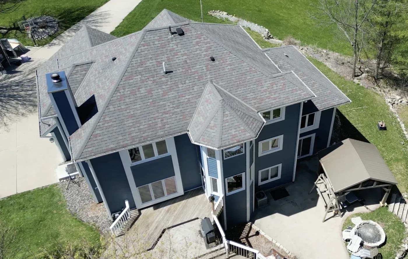 Aerial view of a two-story blue house with gray asphalt shingle roof, featuring a deck, attached garage, and surrounded by green lawn and landscaping
