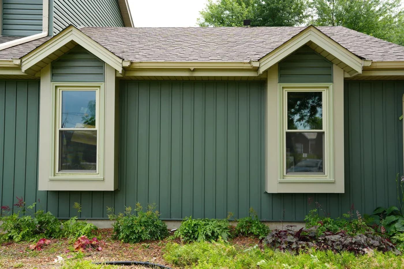 Exterior of a home with green board-and-batten siding, white trim, two windows, and landscaped garden beds.