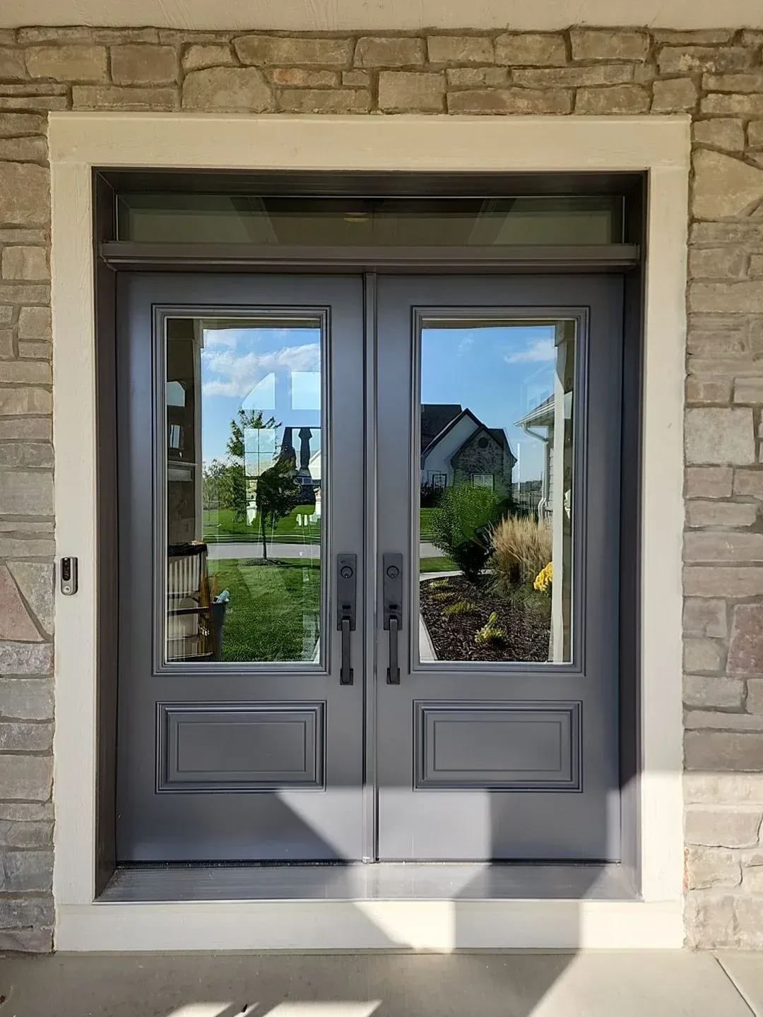 Double black entry doors with glass panes and transom window, set in cream-colored frame within tan brick exterior, looking onto residential driveway and landscaped yard