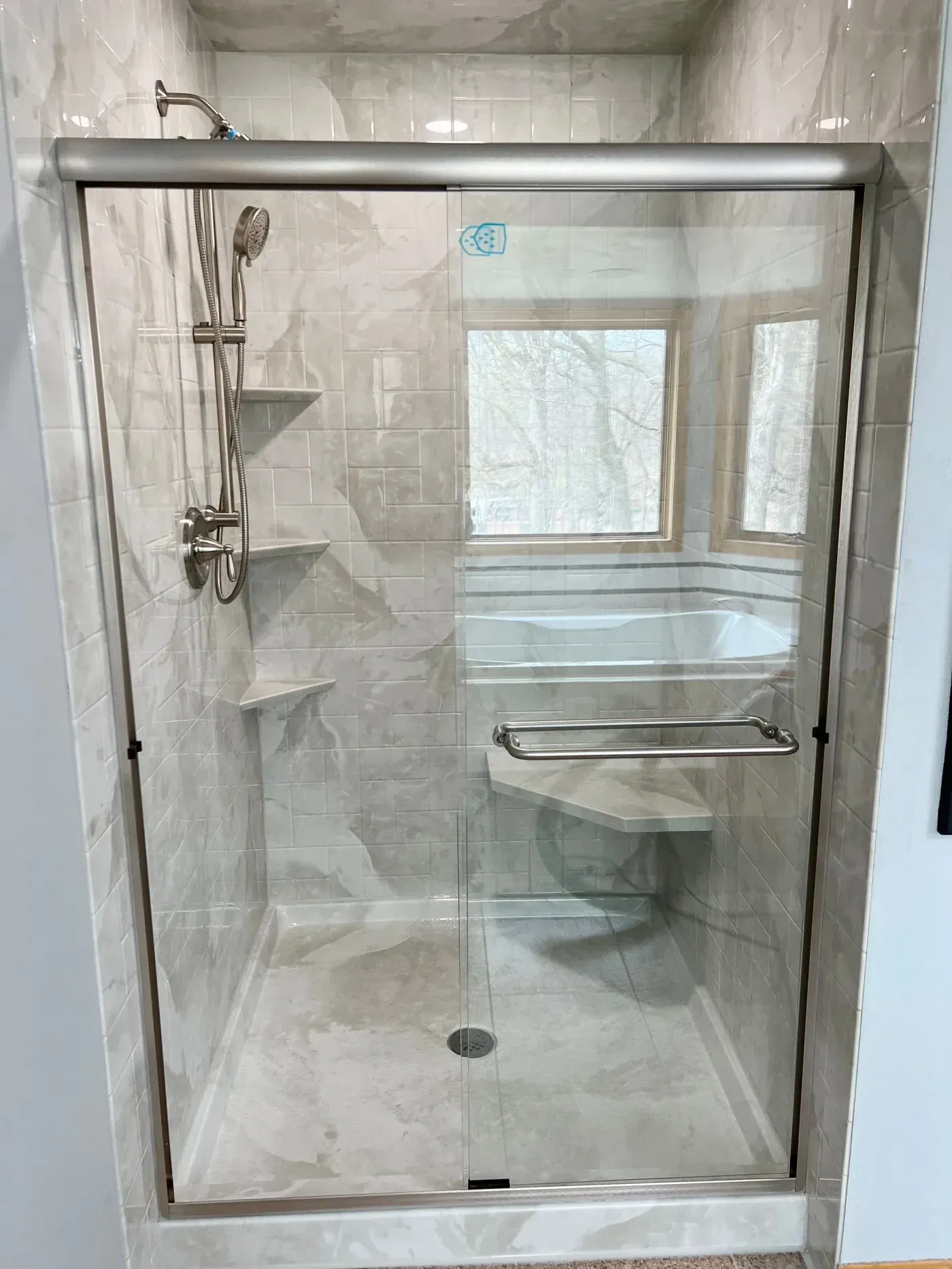 Walk-in shower with marble-look wall panels, glass sliding doors, built-in corner shelves, bench seat, and handheld showerhead in Pewaukee, Wisconsin.