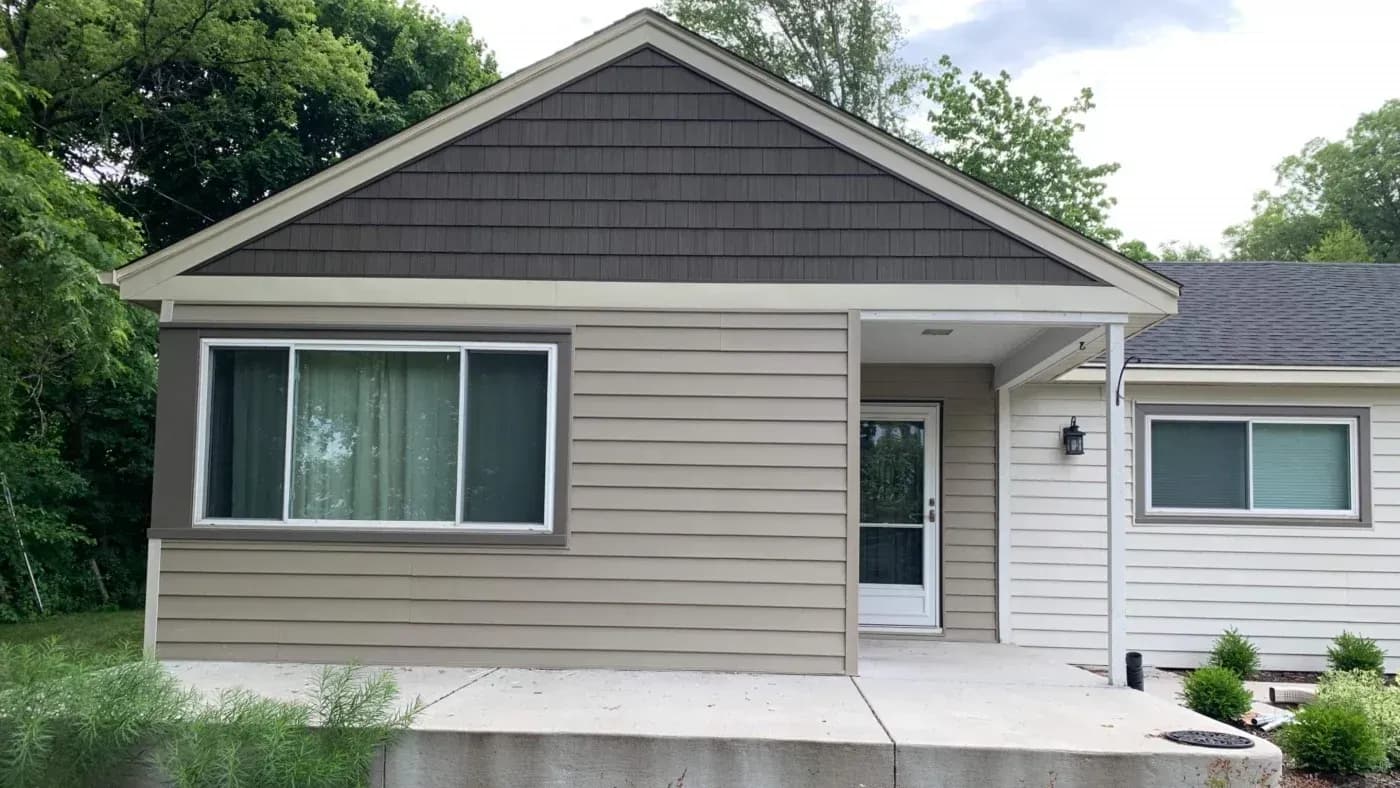Home exterior with tan vinyl siding, dark shake-style gable siding, replacement windows, and new gutters in Menomonee Falls, Wisconsin.