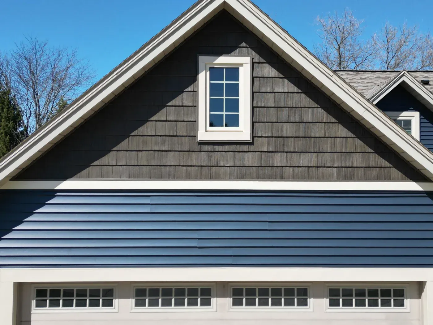 Modern residential home exterior with gabled roofline, charcoal shingle siding, white trim, centered dormer window, and white garage doors with multiple panes