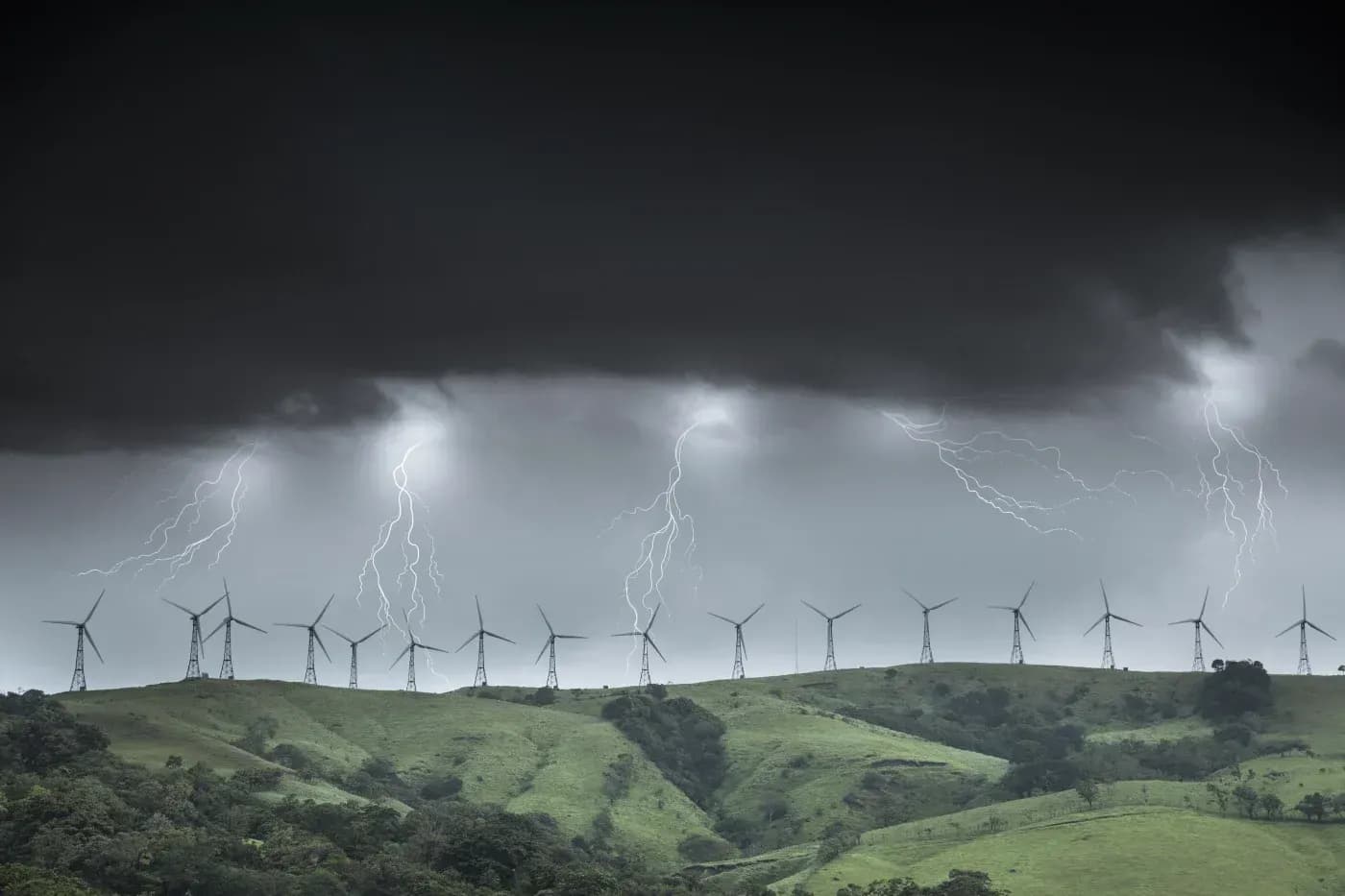 Wind turbines lining green hills beneath dark storm clouds and multiple lightning strikes.