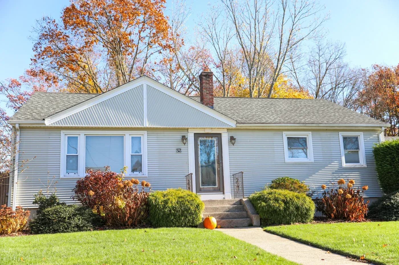 Single-story white vinyl ranch house with metal roof and brick chimney, surrounded by autumn foliage and landscaping, with pumpkin decoration on front lawn