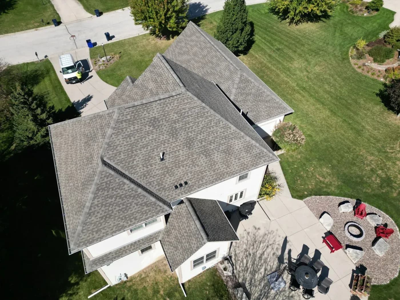 Aerial view of residential home with gray asphalt shingle roofing, white exterior walls, front patio with outdoor furniture and landscaping, driveway with service vehicle, and surrounding lawn and trees