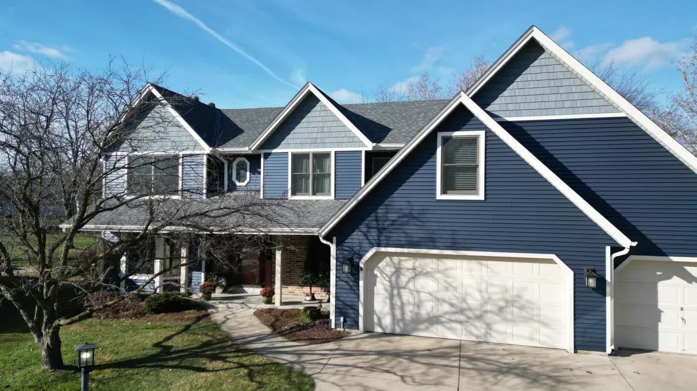 Two-story residential home with blue siding, multiple gabled roofs, white trim, two-car garage, front porch, and bare trees in the yard on a clear day