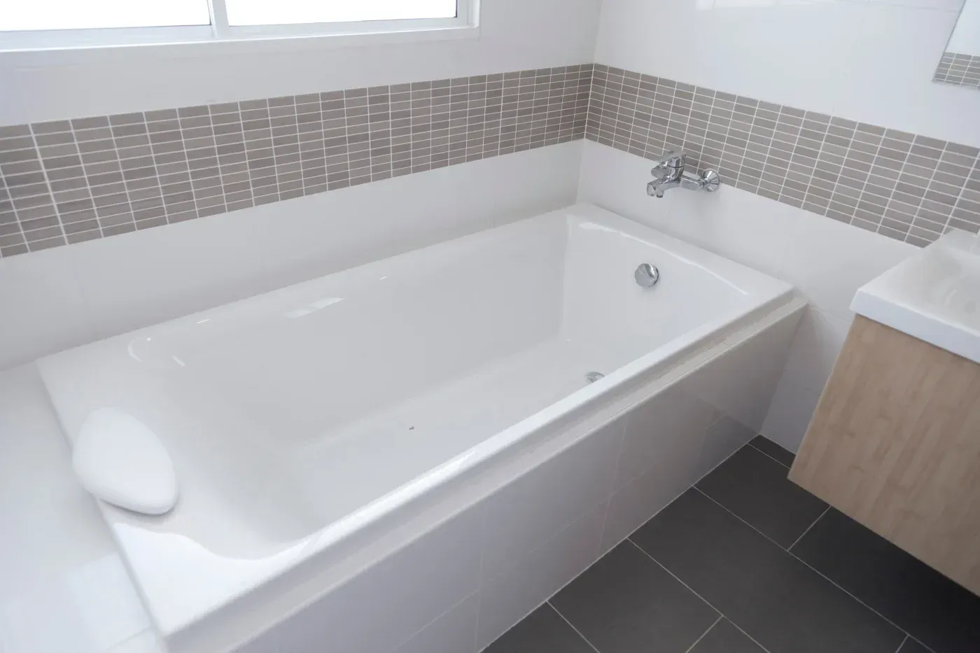 White bathtub beneath a window with gray mosaic tile accents and dark tile flooring.