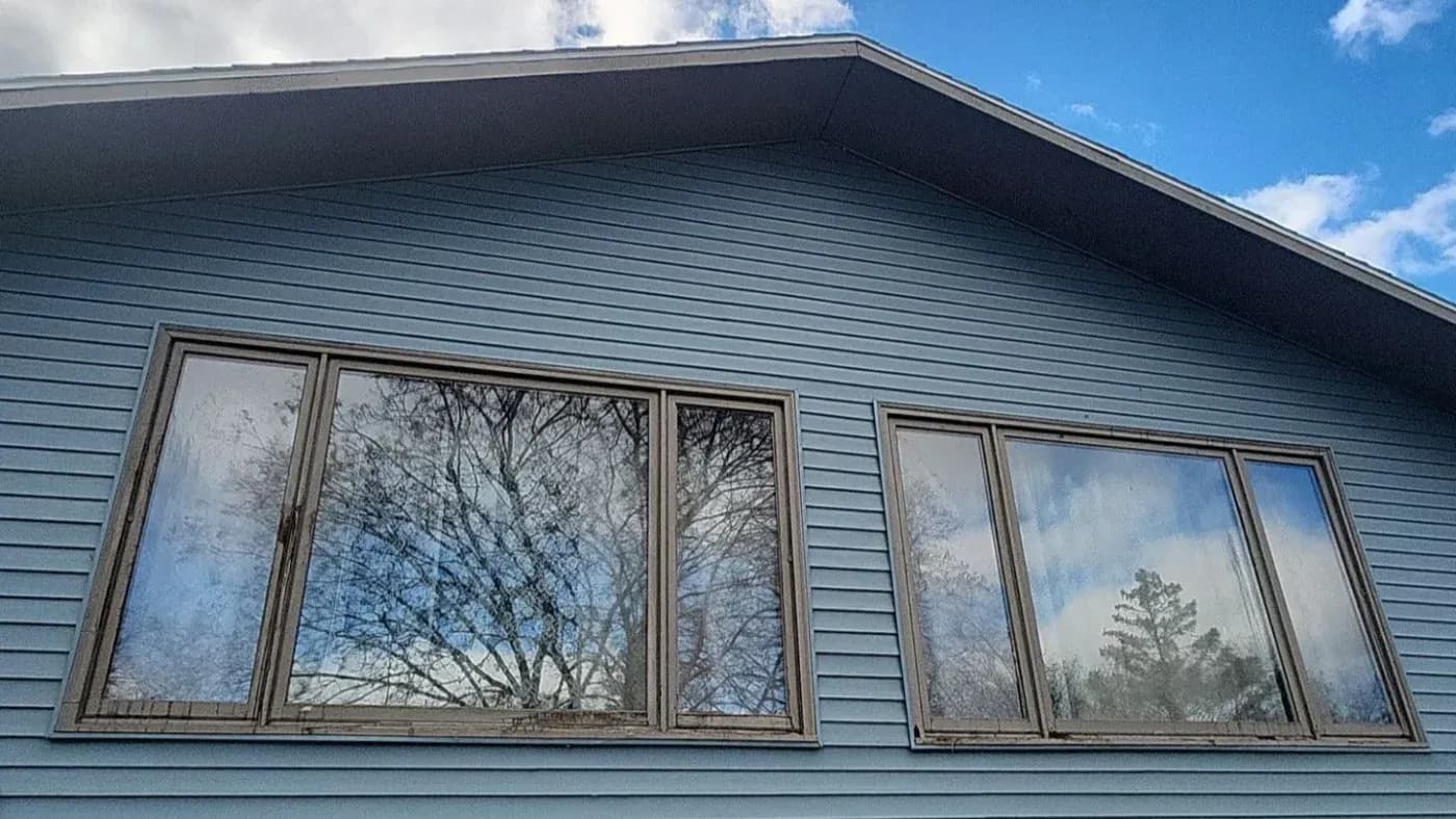 Exterior view of a modern building with blue-gray horizontal siding and two sets of large multi-pane windows reflecting bare trees and sky