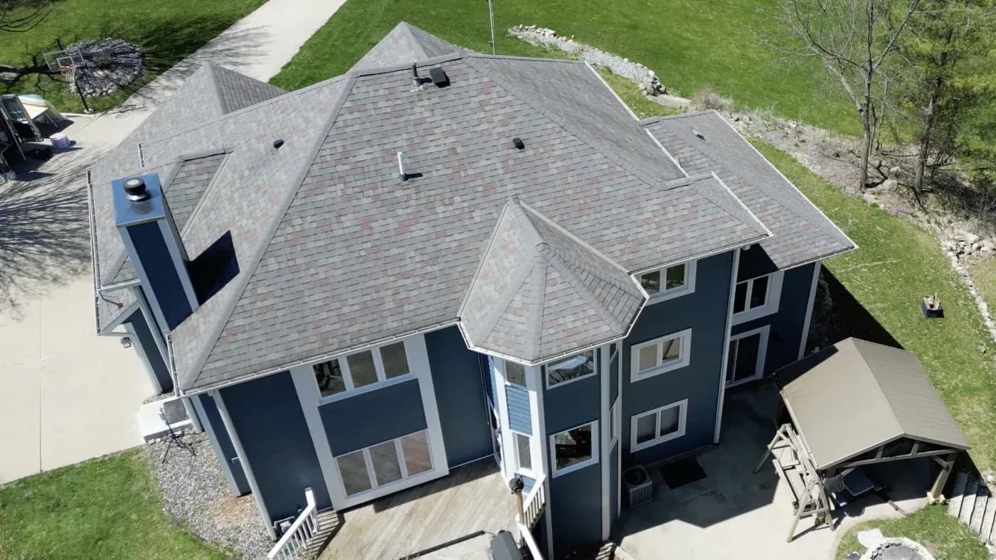 Aerial view of a two-story dark blue house with gray shingle roof, multiple gables, deck, driveway, and surrounding landscaped lawn
