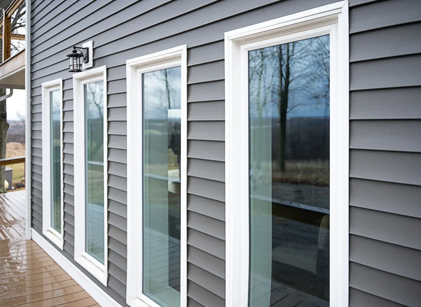 Row of four white-framed sliding glass doors on gray horizontal siding, opening onto a wooden deck with landscape view