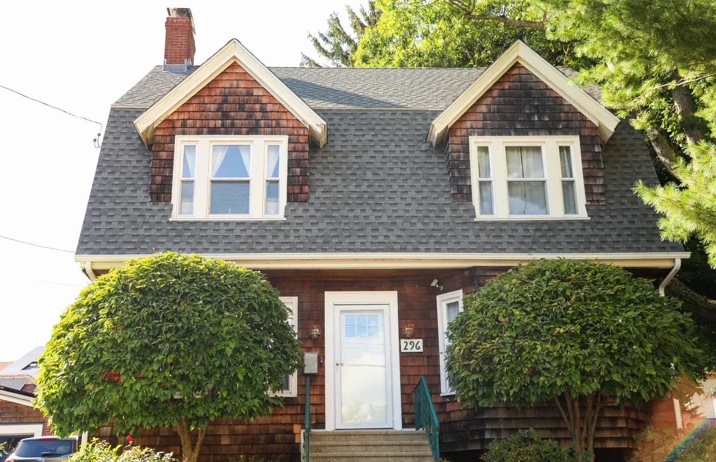 Two-story brick house with dark shingle roof, two dormer windows with white trim, front door numbered 296, flanked by ivy-covered shrubs, surrounded by mature green trees