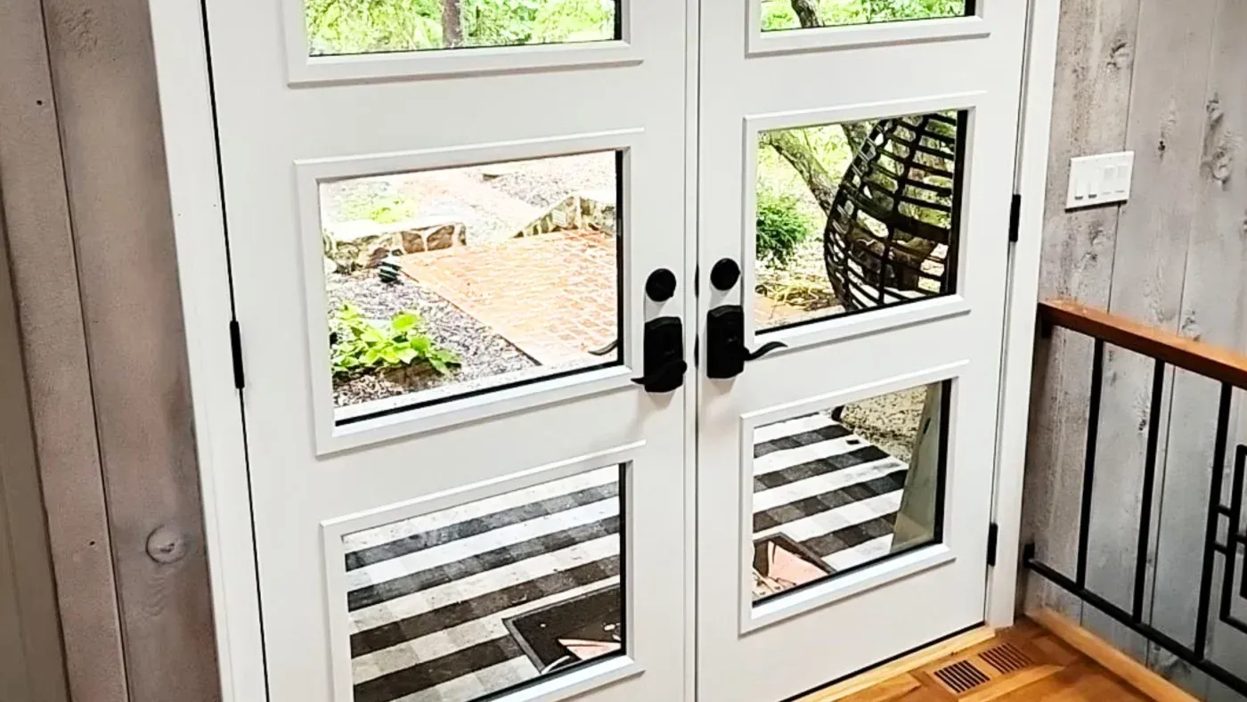 White double French doors with multiple glass panes opening to a backyard patio with brick pavement, landscaping, and wooden pergola structure