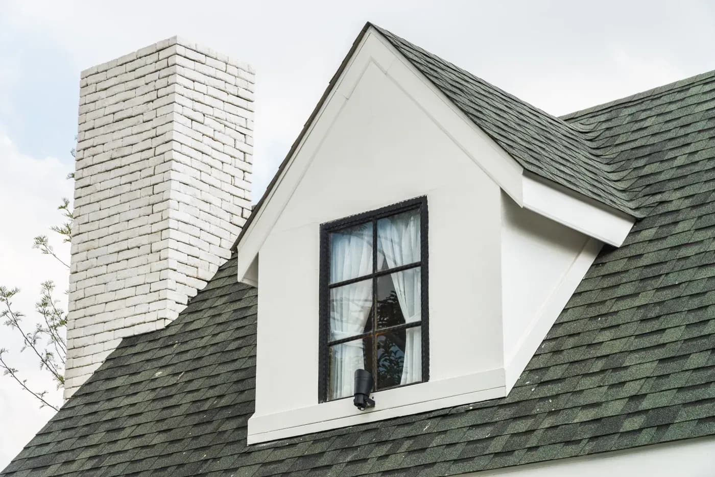 Close-up of a white house dormer with a black-framed window, gray asphalt shingles, and a white brick chimney.