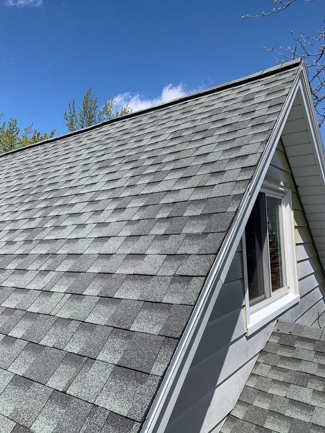 Steep peaked roof with gray asphalt shingles and white trim, featuring a gabled dormer window on a residential house under clear blue sky