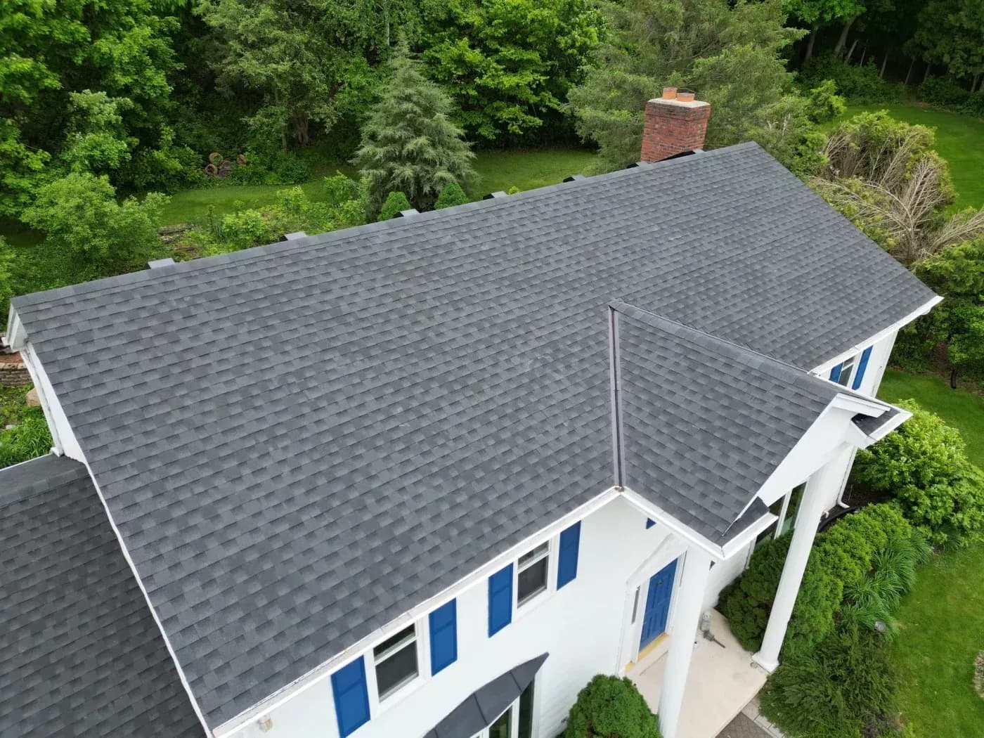 Aerial view of residential house with gray asphalt shingle roof, white walls, blue shutters and doors, red brick chimney, surrounded by green trees and lawn