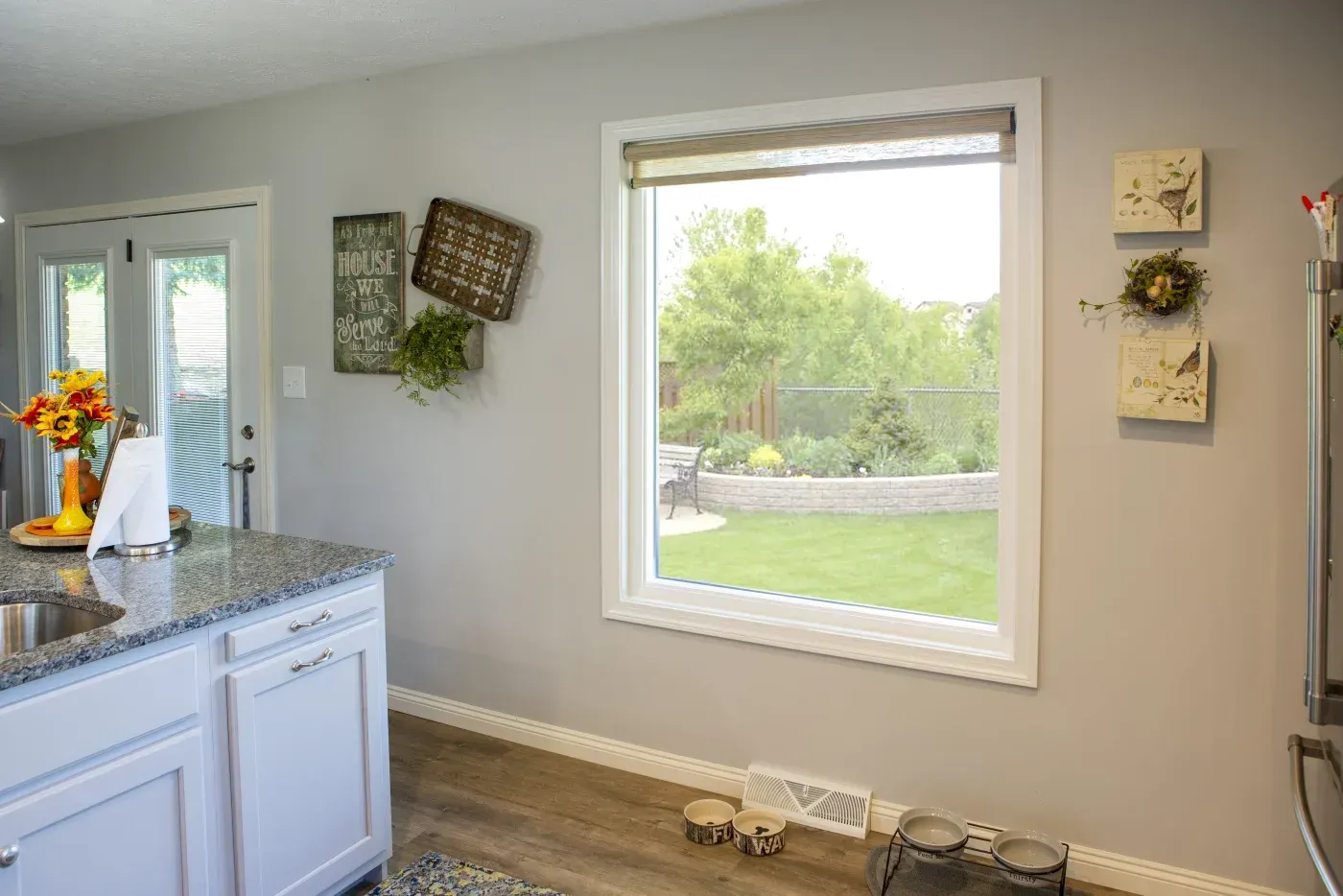 Bright kitchen with white cabinetry and granite countertop overlooking backyard through large window, with sunflower arrangement and wall decor