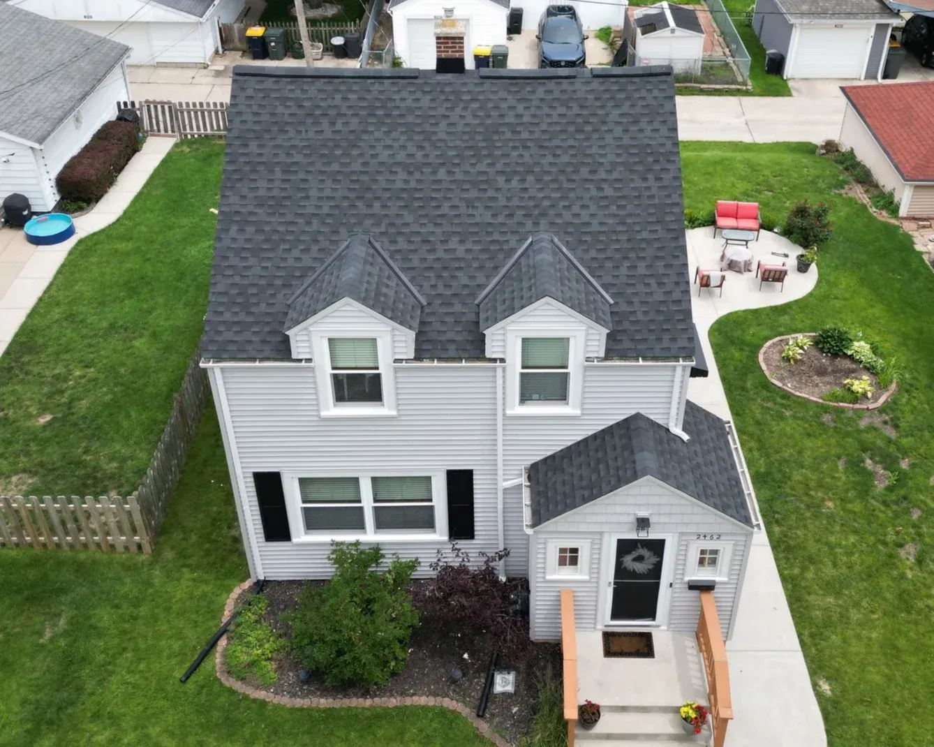 Aerial view of two-story white house with dark gray shingle roof, two front gables, attached garage, front porch, and landscaped yards with neighboring homes visible