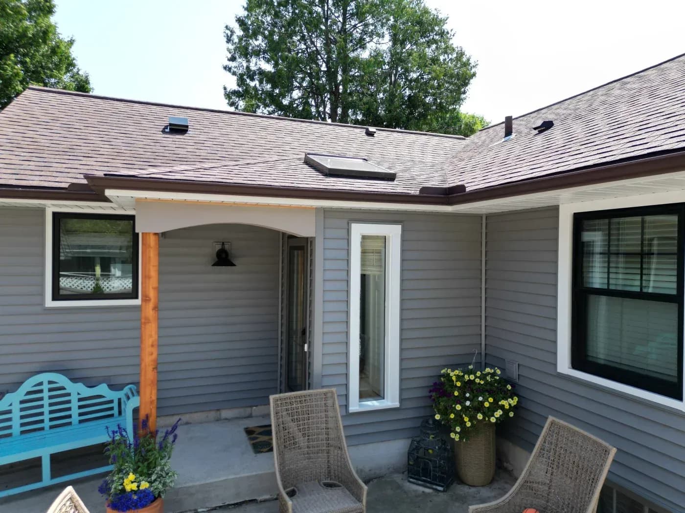 Modern home exterior with covered patio seating area, gray horizontal siding, newly shingled roof with skylights, potted flowers, and wicker chairs