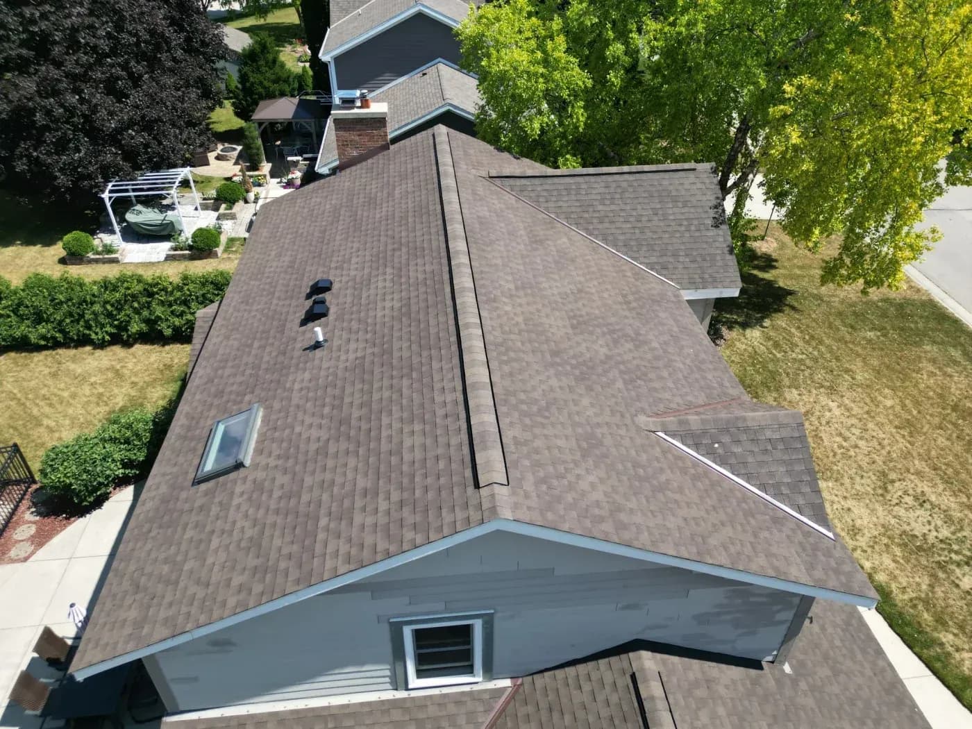 Aerial view of residential house with dark gray shingle roof, skylight, and brick chimney surrounded by green lawn and trees