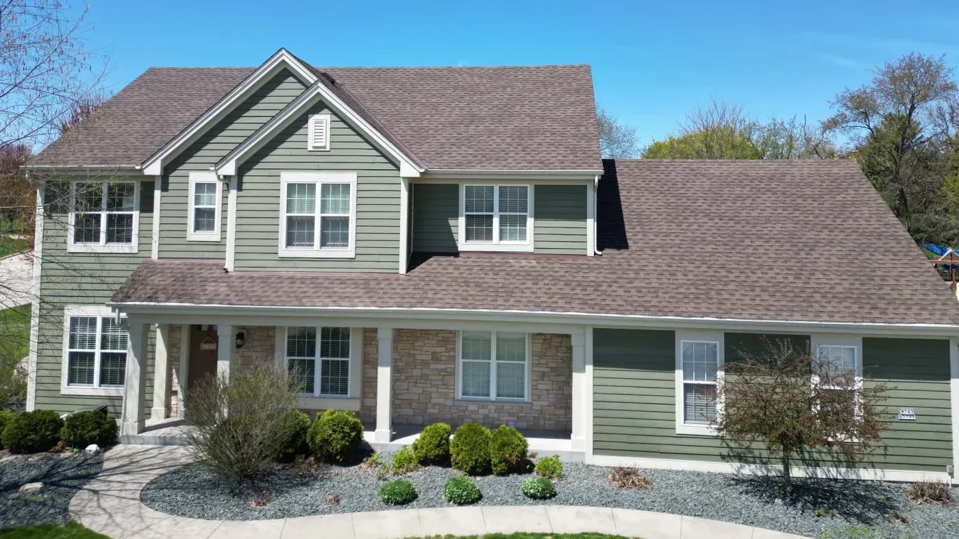 Two-story residential home with green siding, gray shingle roof, stone and siding front facade, multiple windows, covered front porch, and landscaped yard with shrubs and gravel