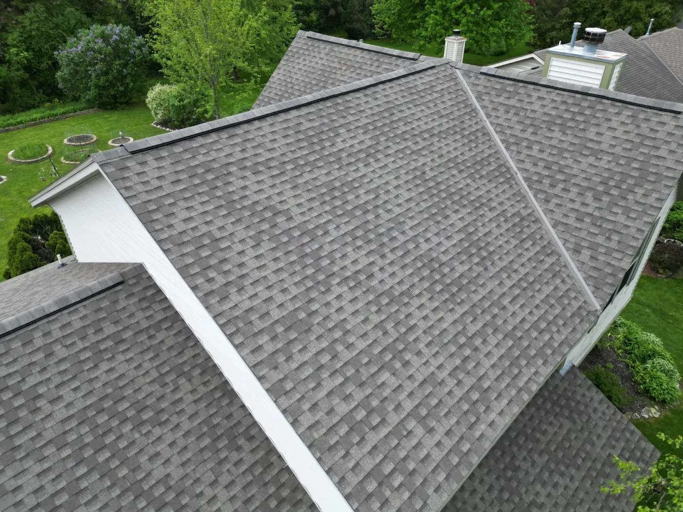 Aerial view of residential roof with gray architectural shingles, white trim, and surrounding landscaped yard with trees and shrubs