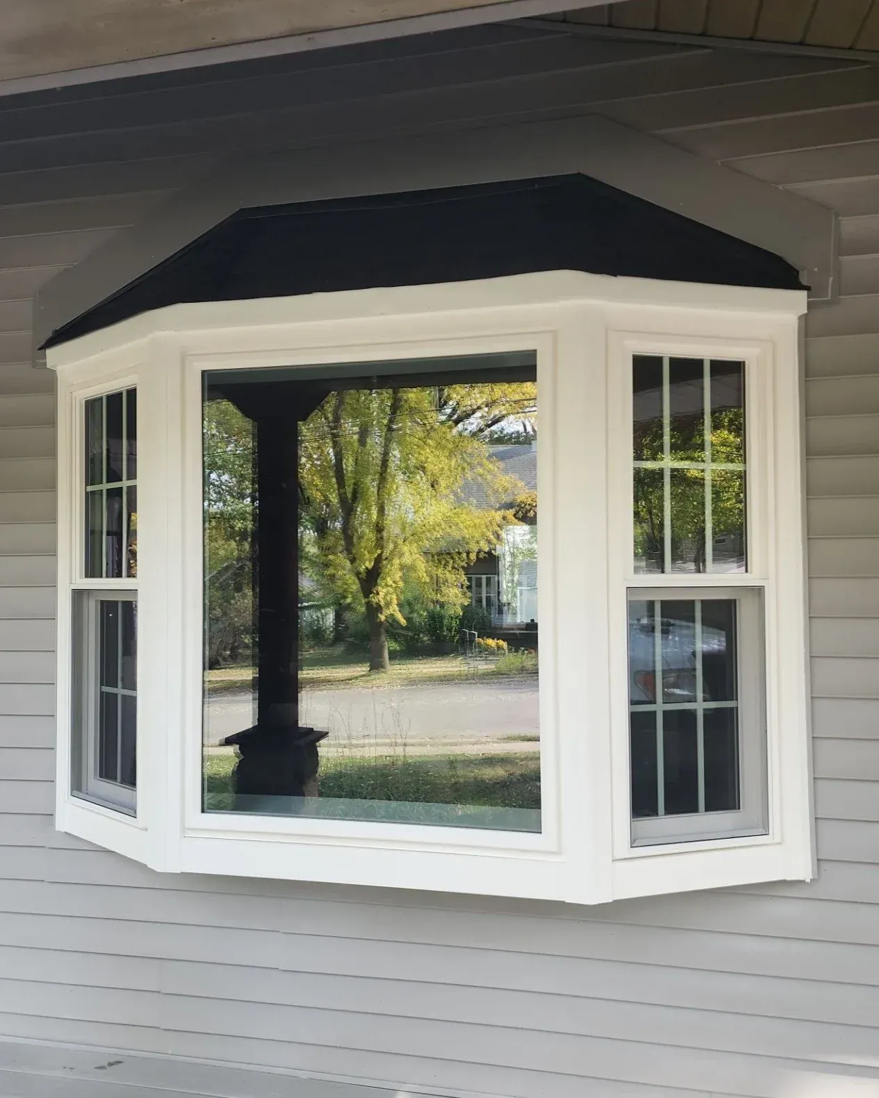 Bay window with cream-colored frame and black roof overhang, featuring multi-pane glass panels reflecting trees and yard view