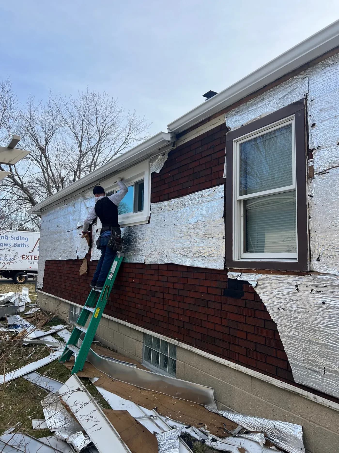 Worker on ladder installing white insulation sheeting over brick exterior wall during home renovation