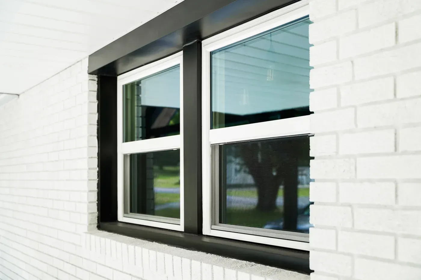 Modern window installation with black aluminum frame and white trim on white brick exterior wall, featuring multiple glass panes with exterior blinds partially drawn