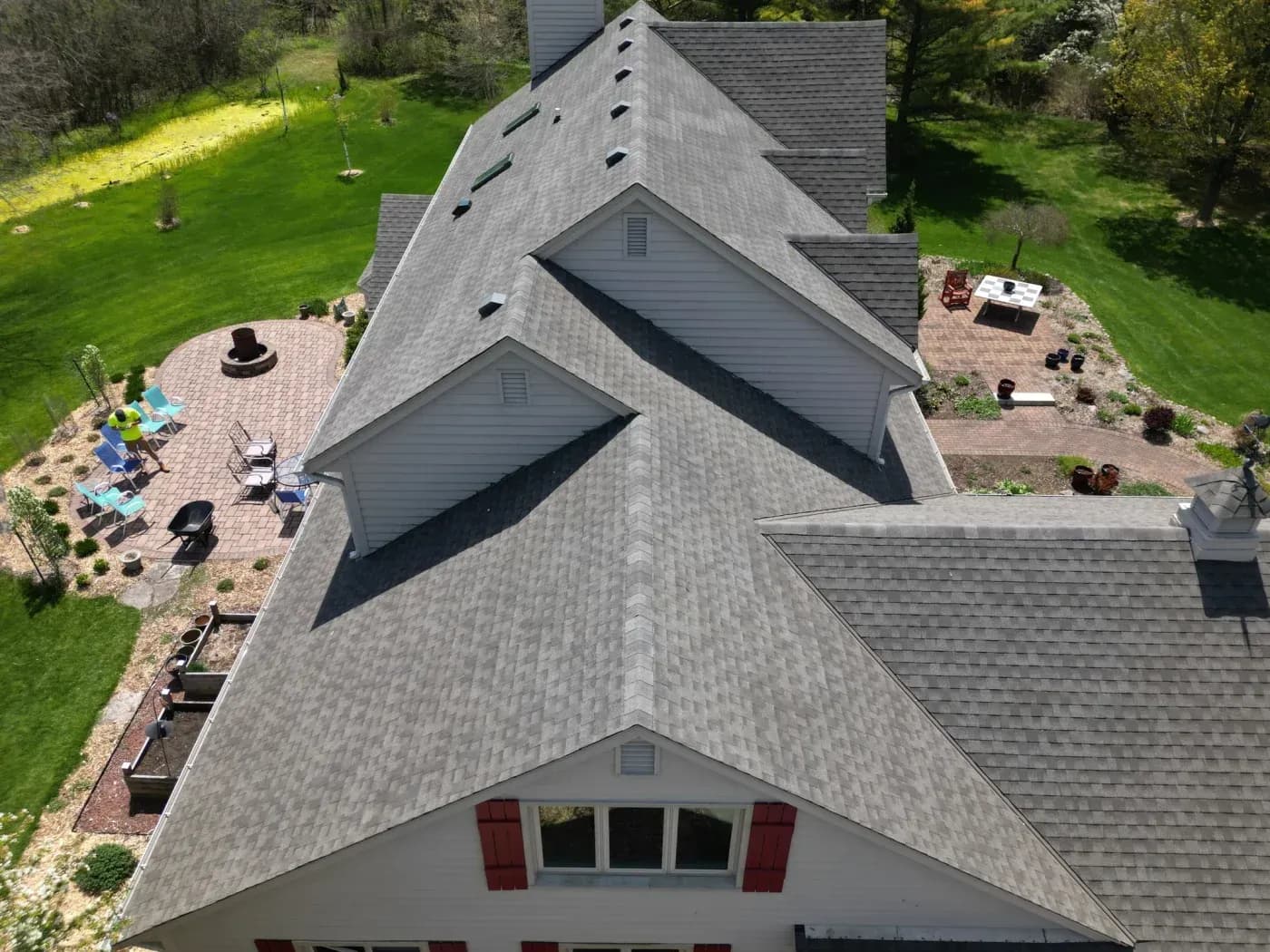 Aerial view of residential home with peaked gray shingled roof, brick patio with lounge chairs and fire pit on left, landscaped yard, and wooded surroundings