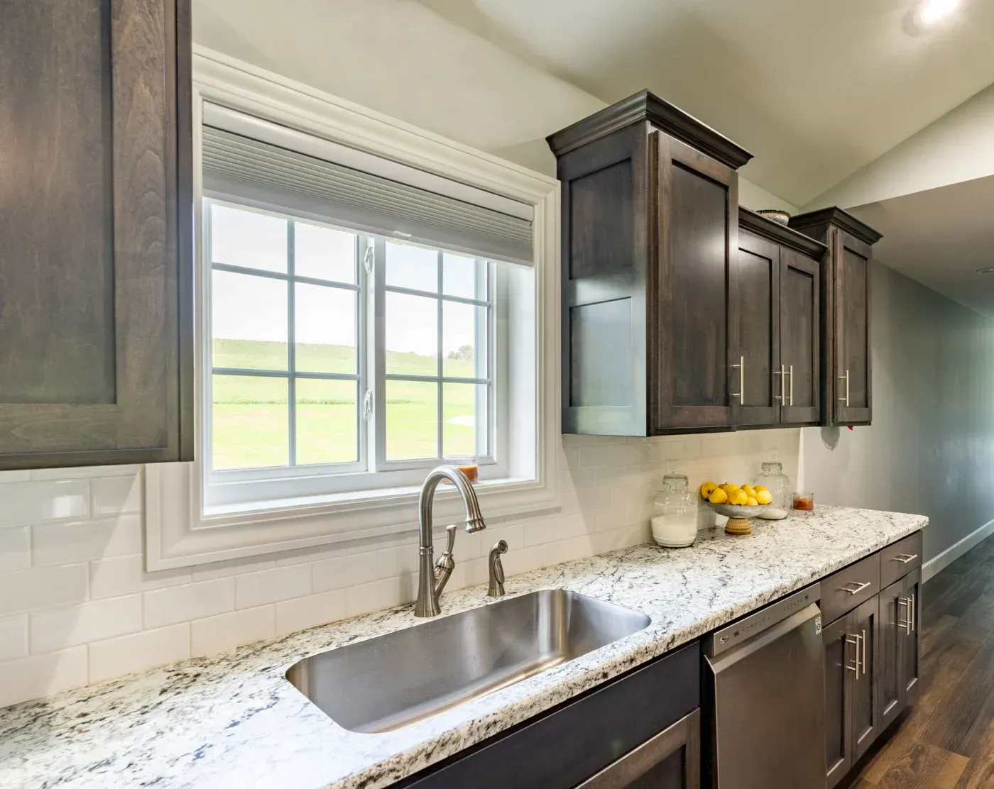 Modern kitchen with stainless steel sink, granite countertop, dark wood cabinets, subway tile backsplash, and large window overlooking green fields