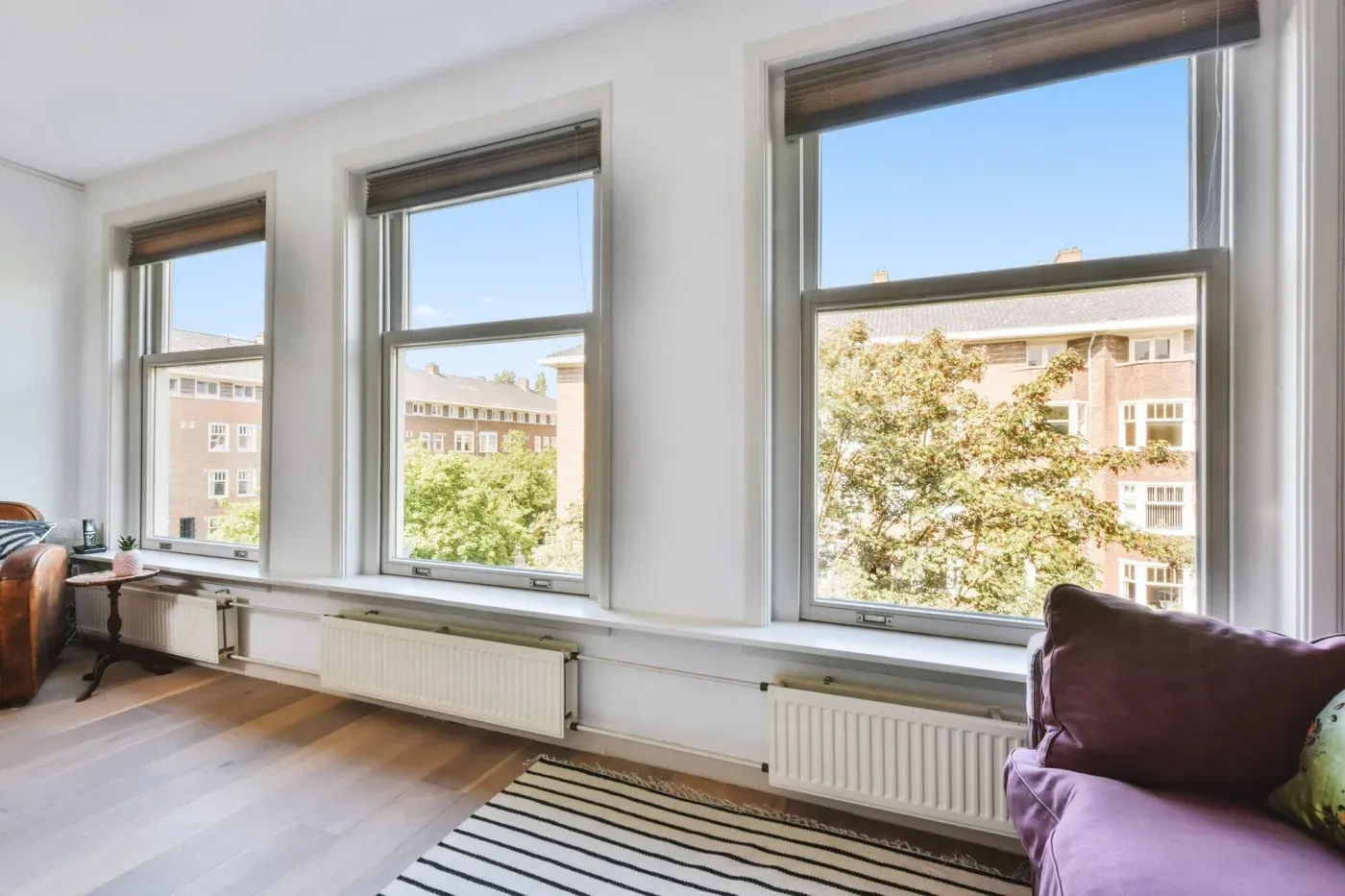 Bright apartment interior with three large windows overlooking a residential neighborhood, white walls, wood flooring, radiators, and purple seating in foreground