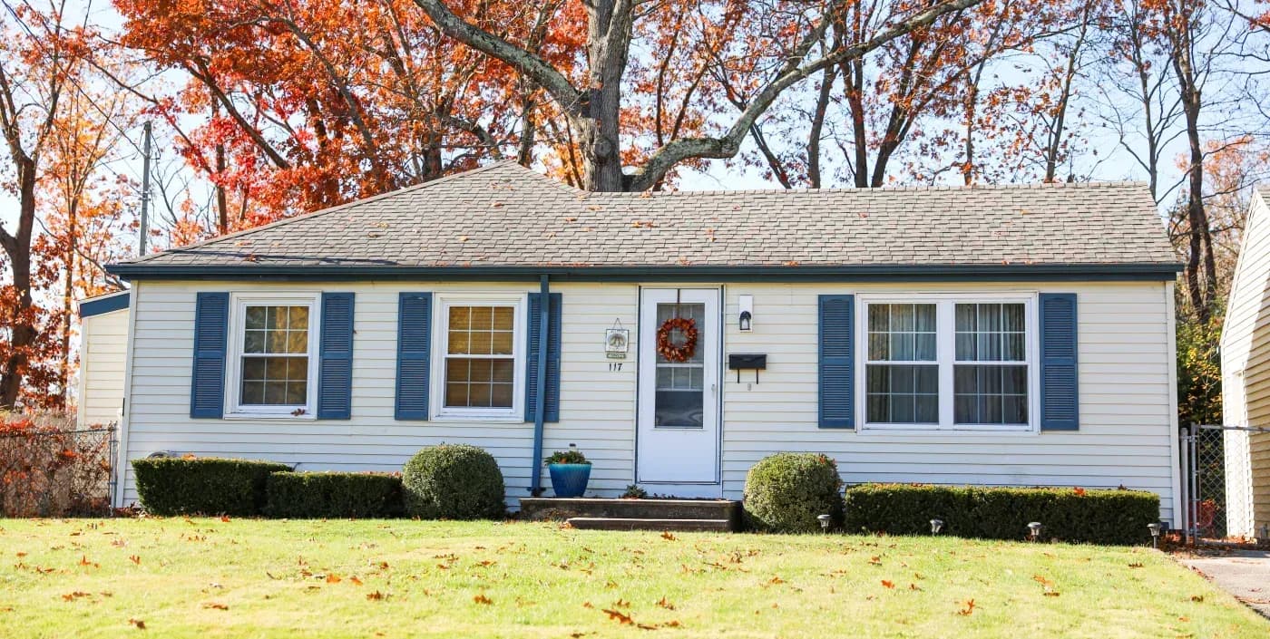 Single-story white ranch house with blue shutters and address 117, featuring a red wreath on the door, surrounded by fall foliage and landscaping
