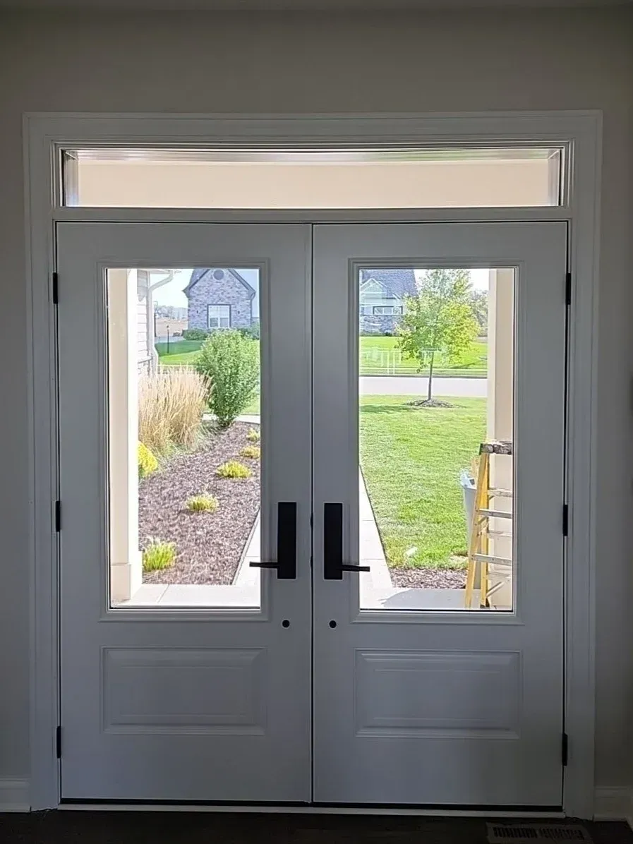 Double glass and panel doors with transom window opening to residential backyard with landscaping and lawn