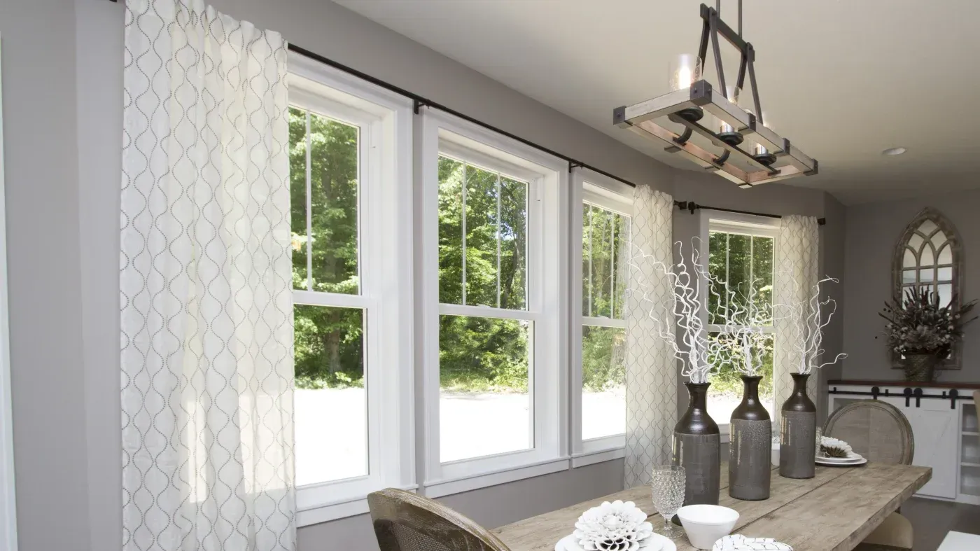 Bright dining room with multiple large windows framed by geometric patterned curtains, overlooking green trees, featuring a wooden table with decorative dark vases and white flowers, metal pendant lighting, and gray walls