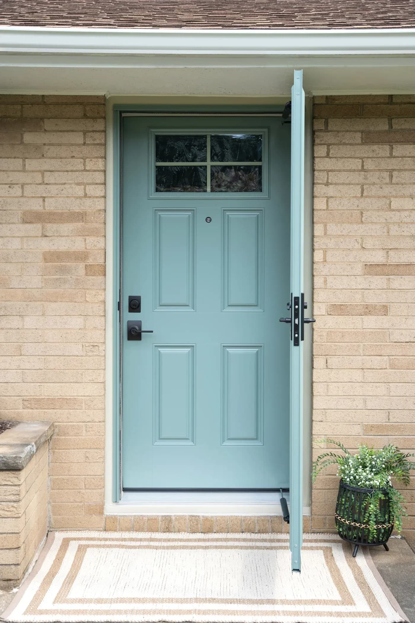 Light blue front door with six panels and window transom on tan brick house, flanked by white trim and potted plant on right side of entryway