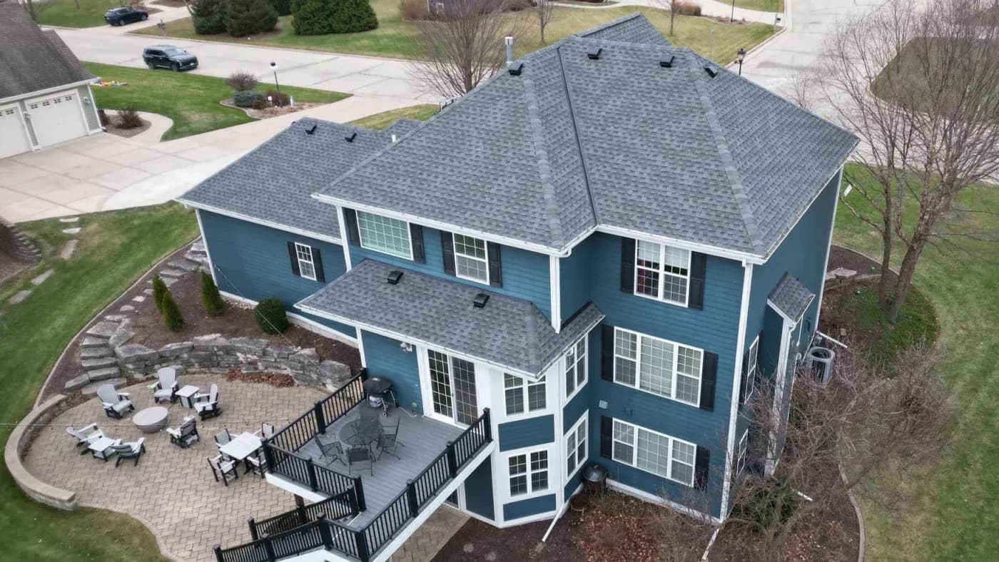 Aerial view of a two-story teal and white house with gray shingle roof, rear deck, and curved paver patio with outdoor seating in a residential neighborhood