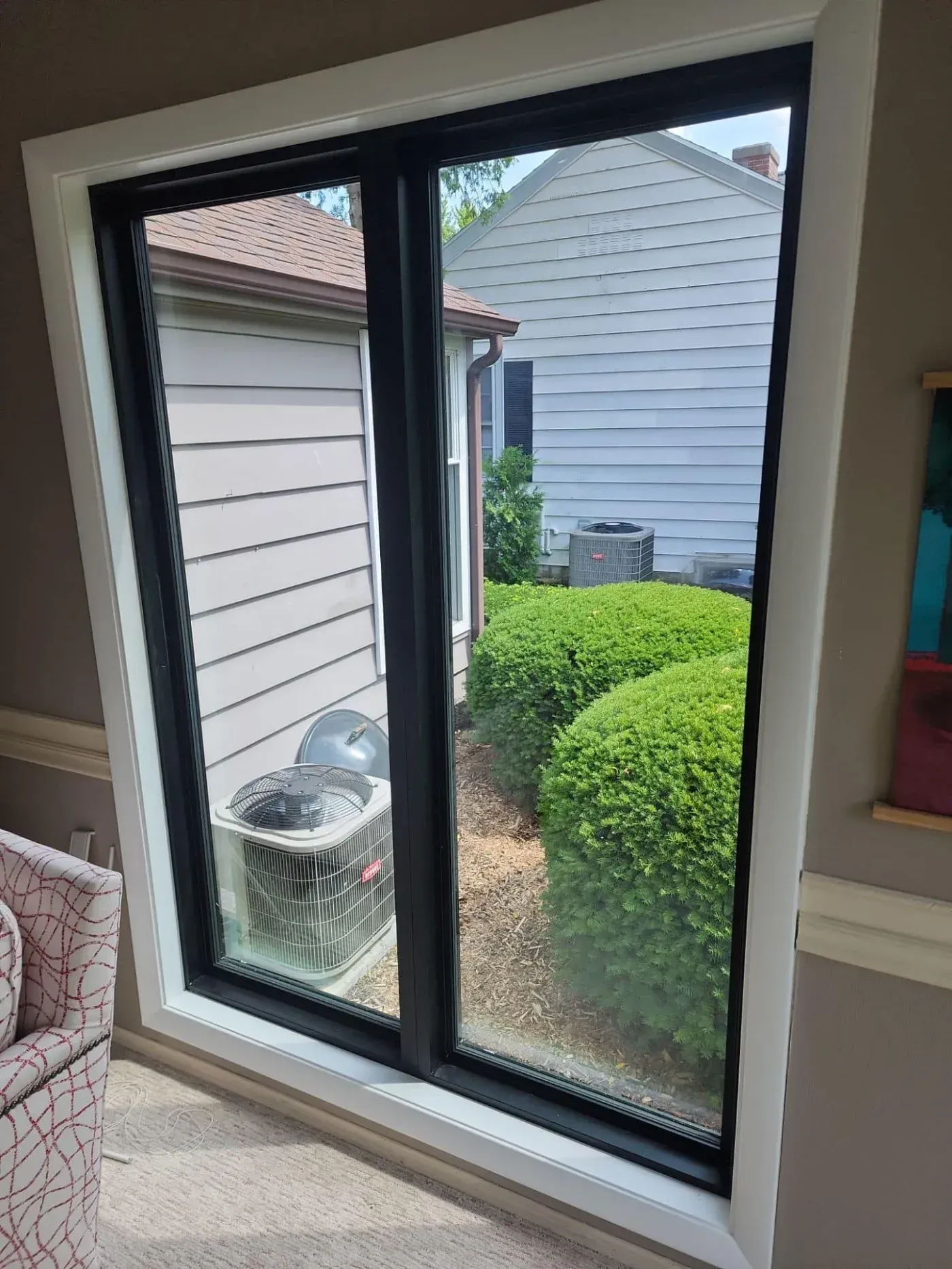 Sliding glass door with black frame opens to backyard view showing air conditioning unit, green hedge, and neighboring houses with siding