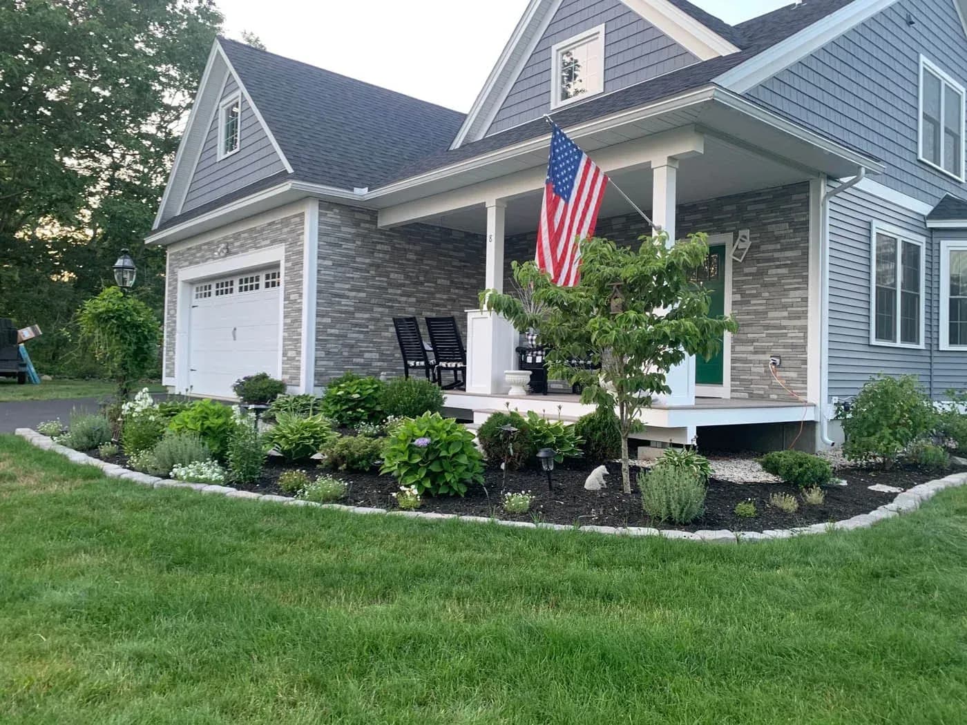Modern two-story house with brick and siding exterior, front porch with American flag, white garage, manicured landscaping with shrubs and plants, and green lawn