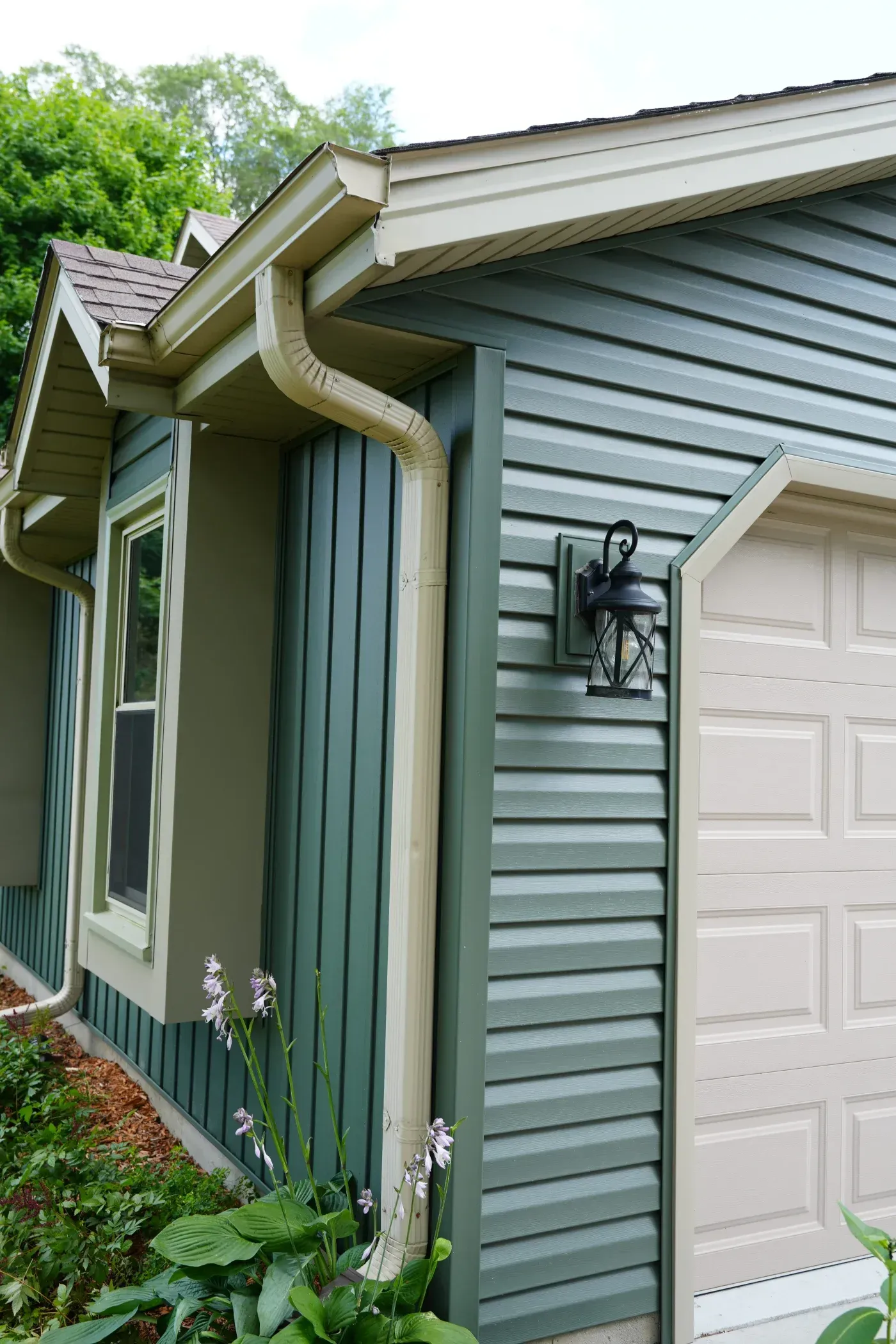 Modern house exterior with green and white siding, cream-colored gutters and downspout, white garage door, and landscape plants with purple flowers at foundation