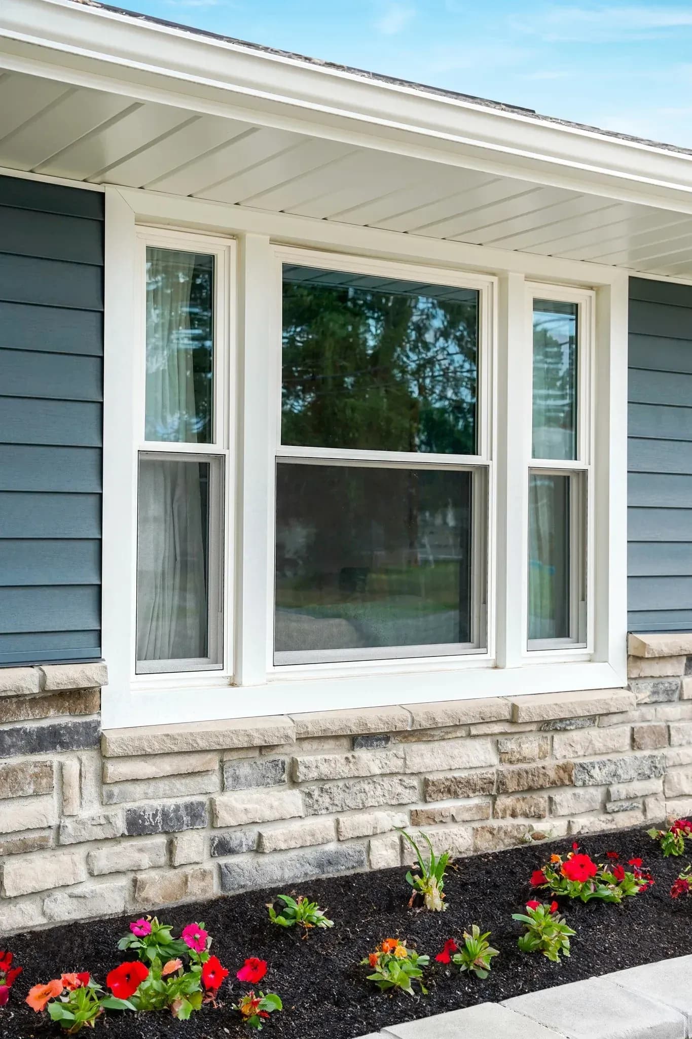 Home exterior showing white-framed double-hung windows with stone foundation, covered porch, and flower garden bed below