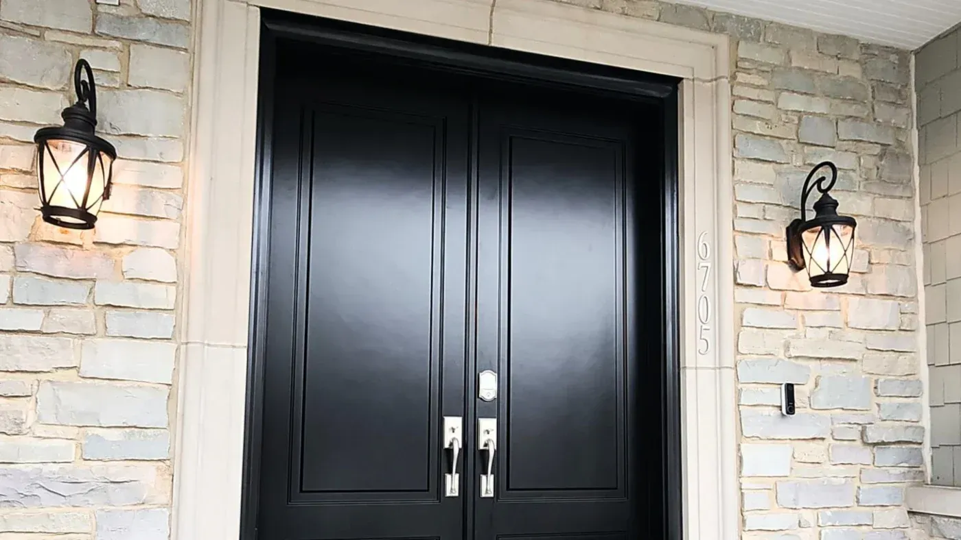 Black double entry doors with chrome handles flanked by lit lantern sconces on a cream brick exterior