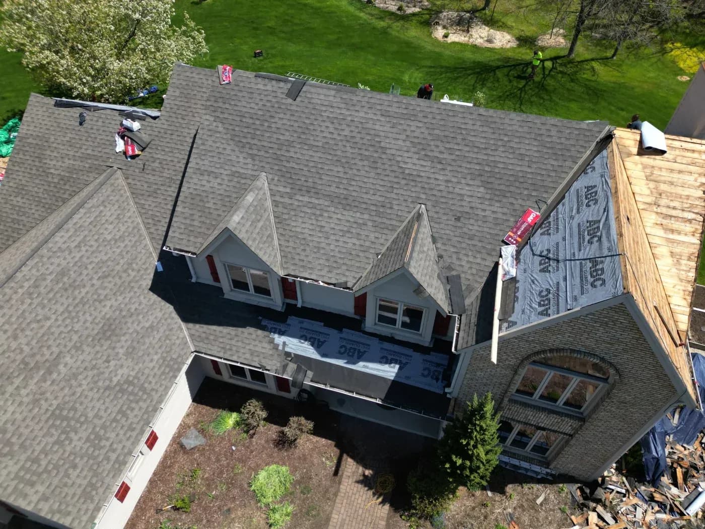 Aerial view of a house roof under construction with new asphalt shingles being installed, workers on the roof, and protective underlayment visible on sections being worked on