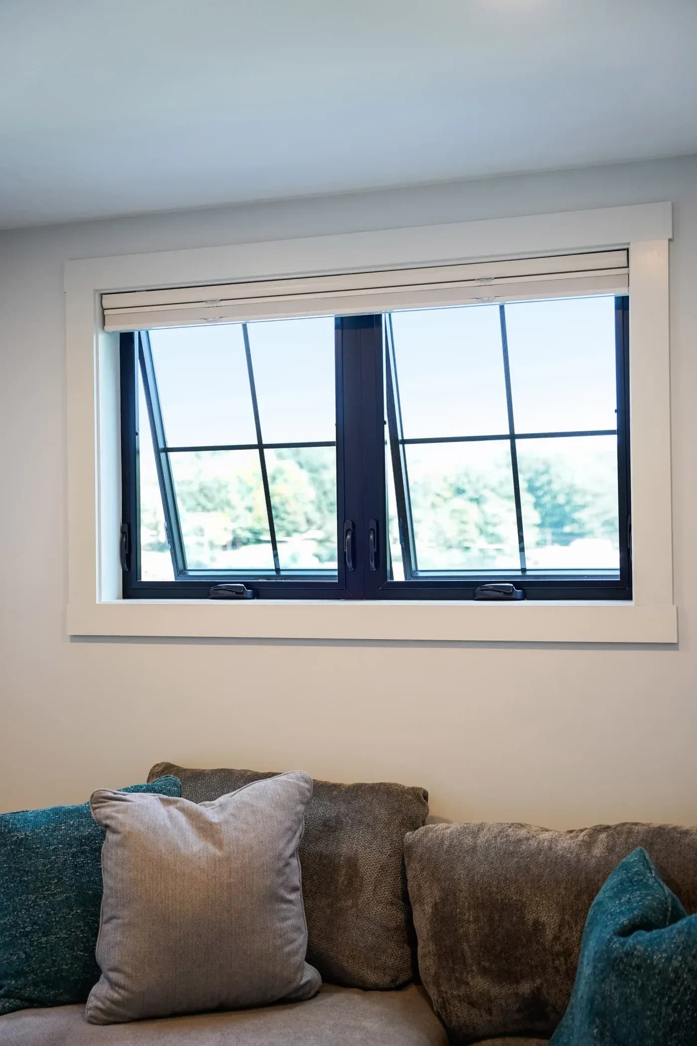 Large horizontal window with dark frame and grids above a beige sofa with teal and gray pillows, overlooking trees and sky