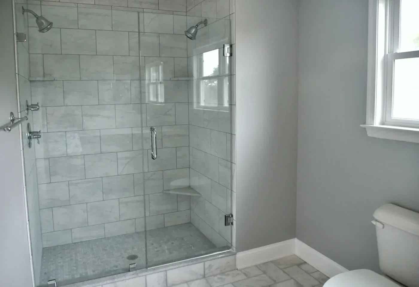 Glass-enclosed walk-in shower with marble-look tile, dual showerheads, and built-in corner shelves.