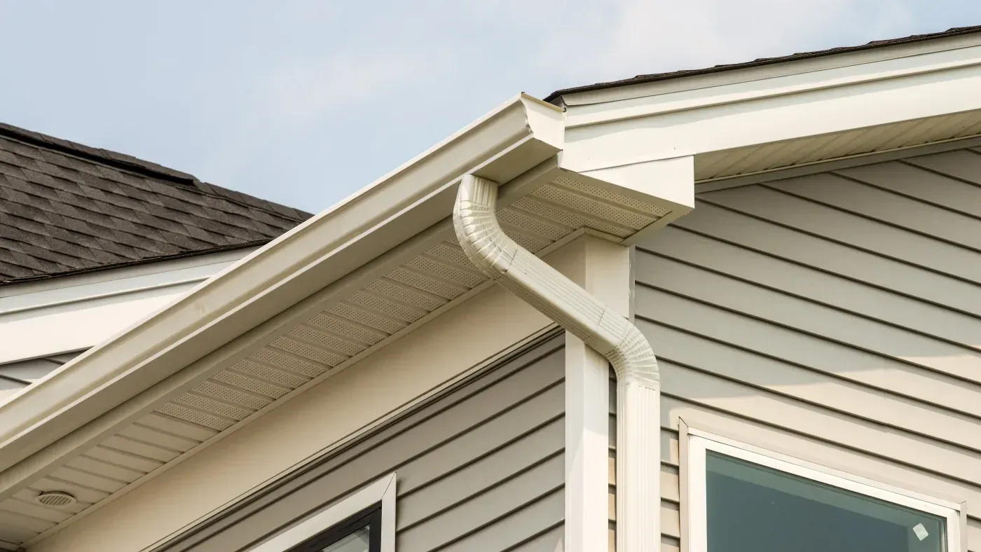 White vinyl gutters and downspout directing water away from home's exterior with light gray siding and dark shingle roof
