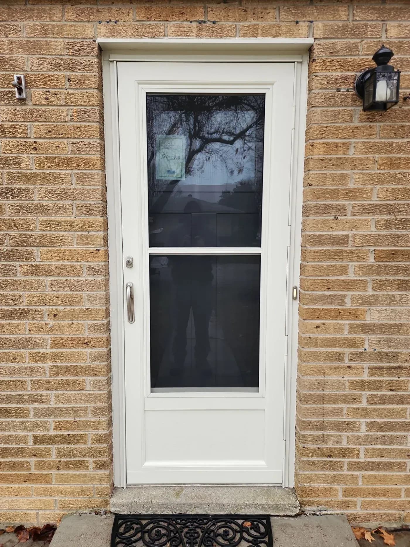 White exterior door with screen and dark glass panels set in tan brick wall, flanked by wall-mounted lantern and doorbell